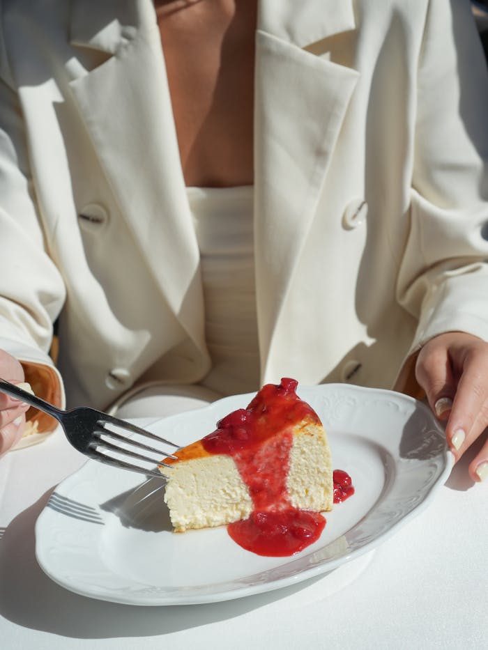 A stylish person in white enjoys a cheesecake topped with strawberry sauce.