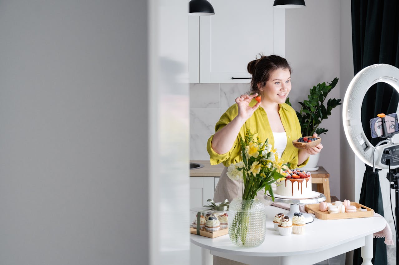 Delighted young female influencer in casual clothes smiling and demonstrating fresh berries while decorating appetizing cake during recording vlog on smartphone in kitchen